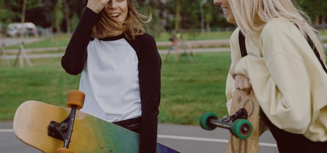 A teenager holding a yellow and blue longboard talking to her friend